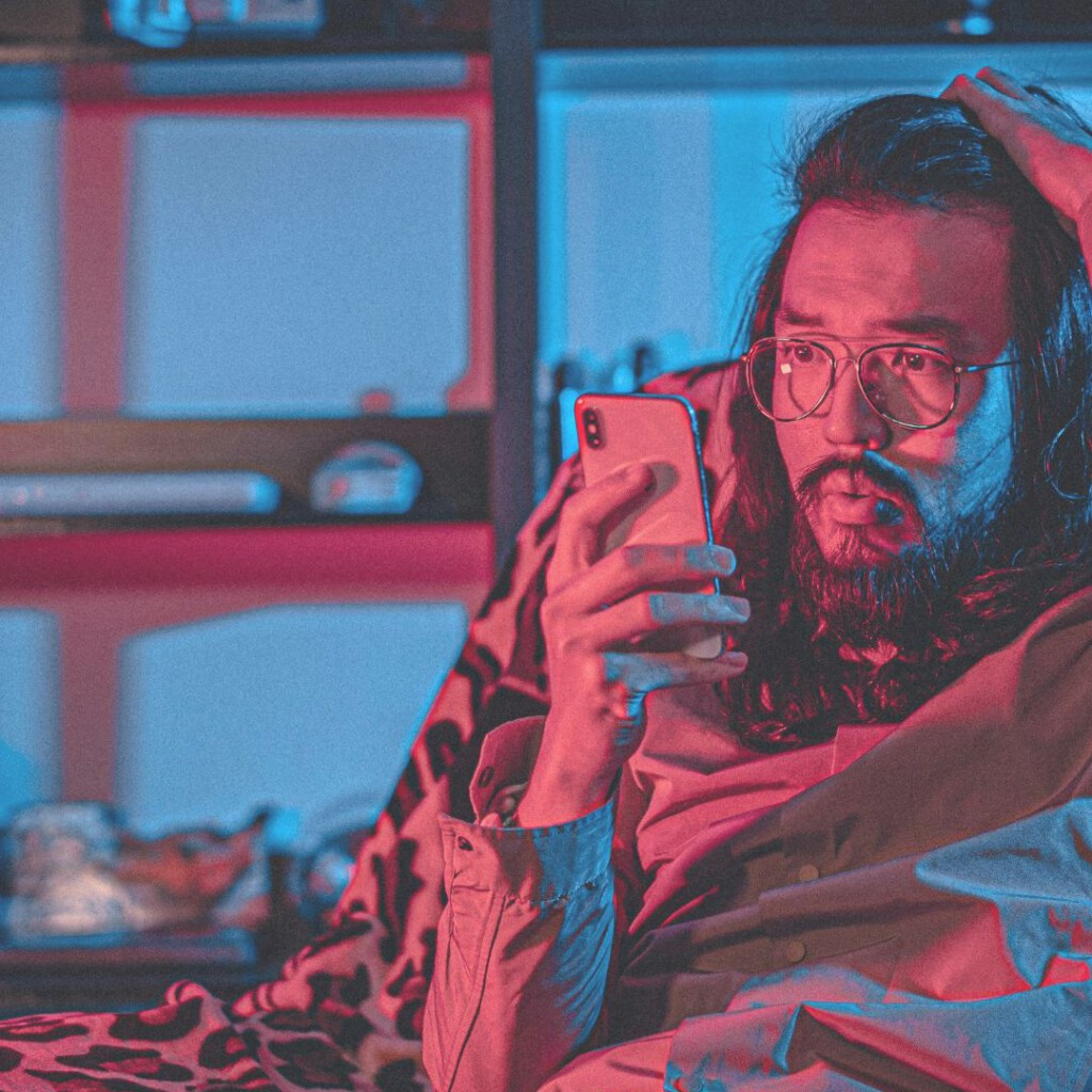 Ein langhaariger Mann mit Brille und Bart schaut besorgt auf sein Smartphone, Hand am Kopf.