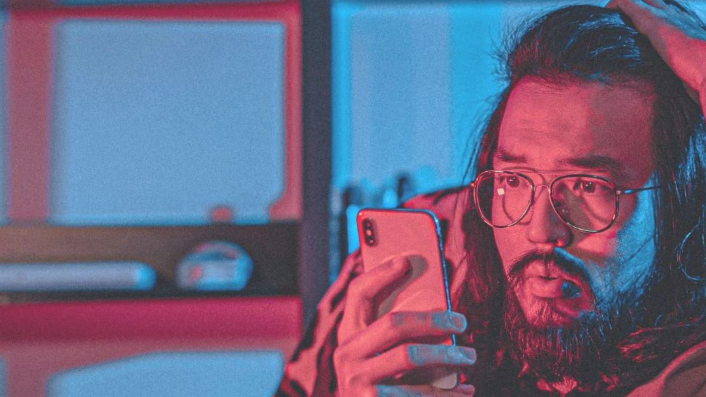 Ein langhaariger Mann mit Brille und Bart schaut besorgt auf sein Smartphone, Hand am Kopf.