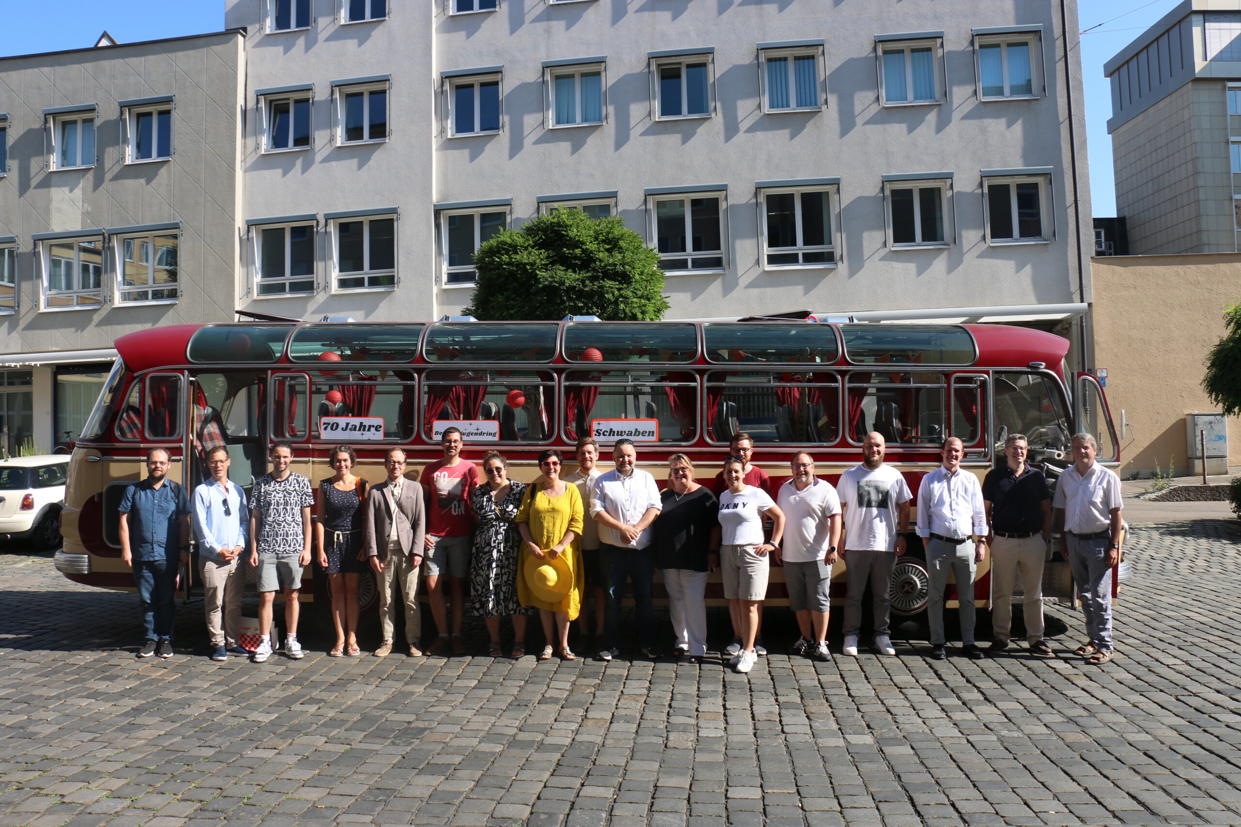 Achtzehn Menschen stehen aufgereiht nebeneinander vor einem rot-beigen Oldtimer-Bus und lachen in die Kamera