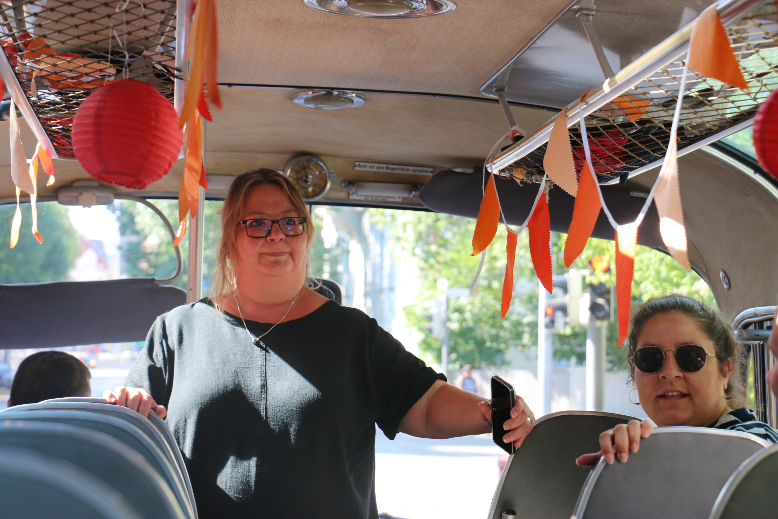 Eine blonde Frau mit Brille im schwarzen Shirt steht im Gang eines Oldtimer-Buses der innen mit orangenen Girlanden und roten Lampions dekoriert ist. Daneben sitzt eine Frau mit Sonnenbrille und braunen Haaren.