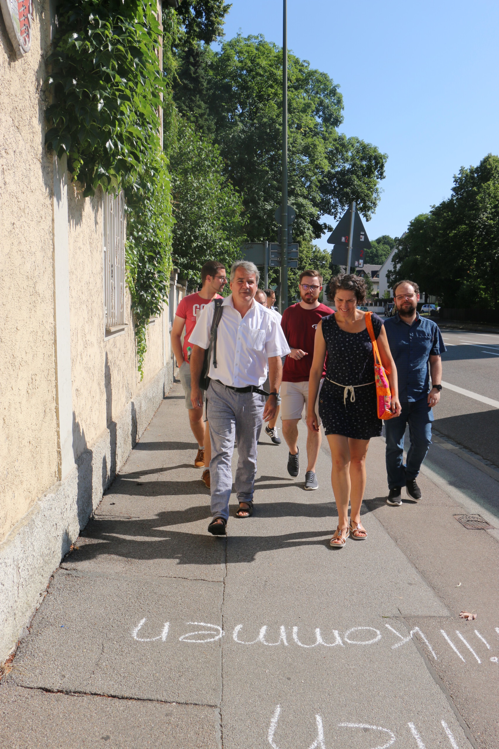 Fünf Menschen laufen in der Sonne auf einem breiten Gehweg entlang einer bewachsenen Hauswand