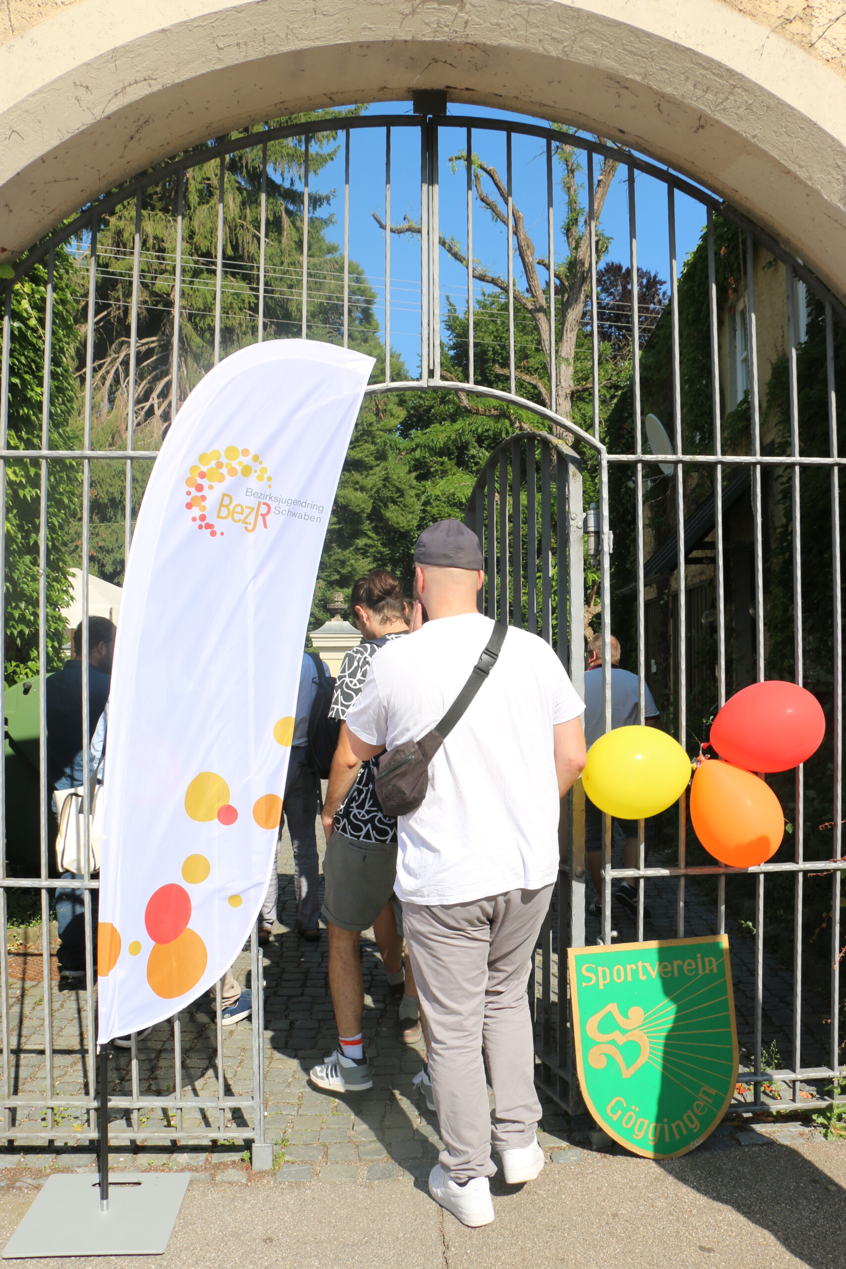 Durch ein Eisentor geht ein Mann mit weißem Shirt, Cap und schwarzer Umhängetasche. Das Tor ist mit orangenen, roten und gelben Luftballons, einer Beachflag vom BezJR und einem grünen Wappen vom Sportverein Göggingen geschmückt