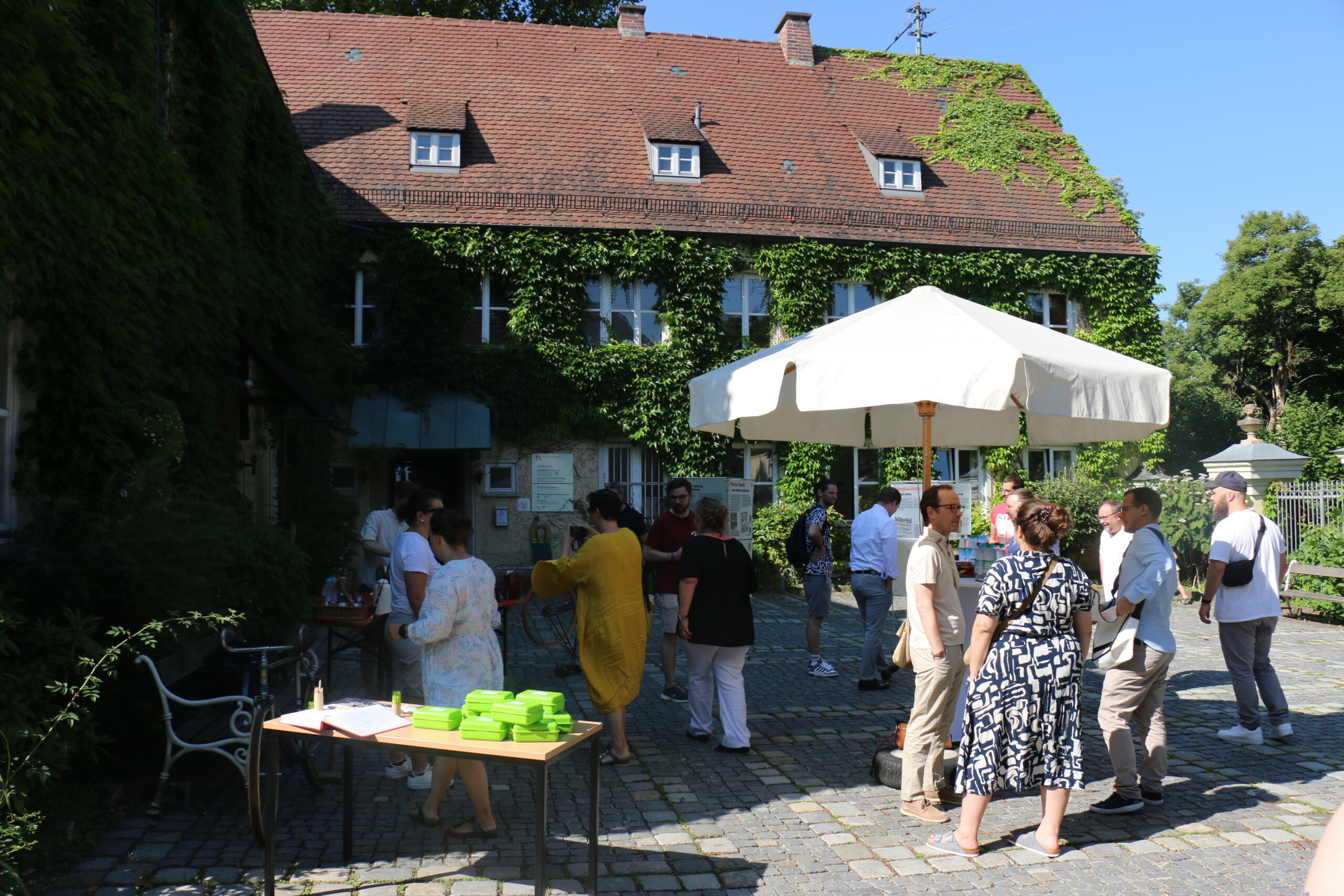 In einem gepflasterten Innenhof mit weißem Sonnenschirm vor einem grün bewachsenen Haus stehen viele Personen locker beieinander und unterhalten sich