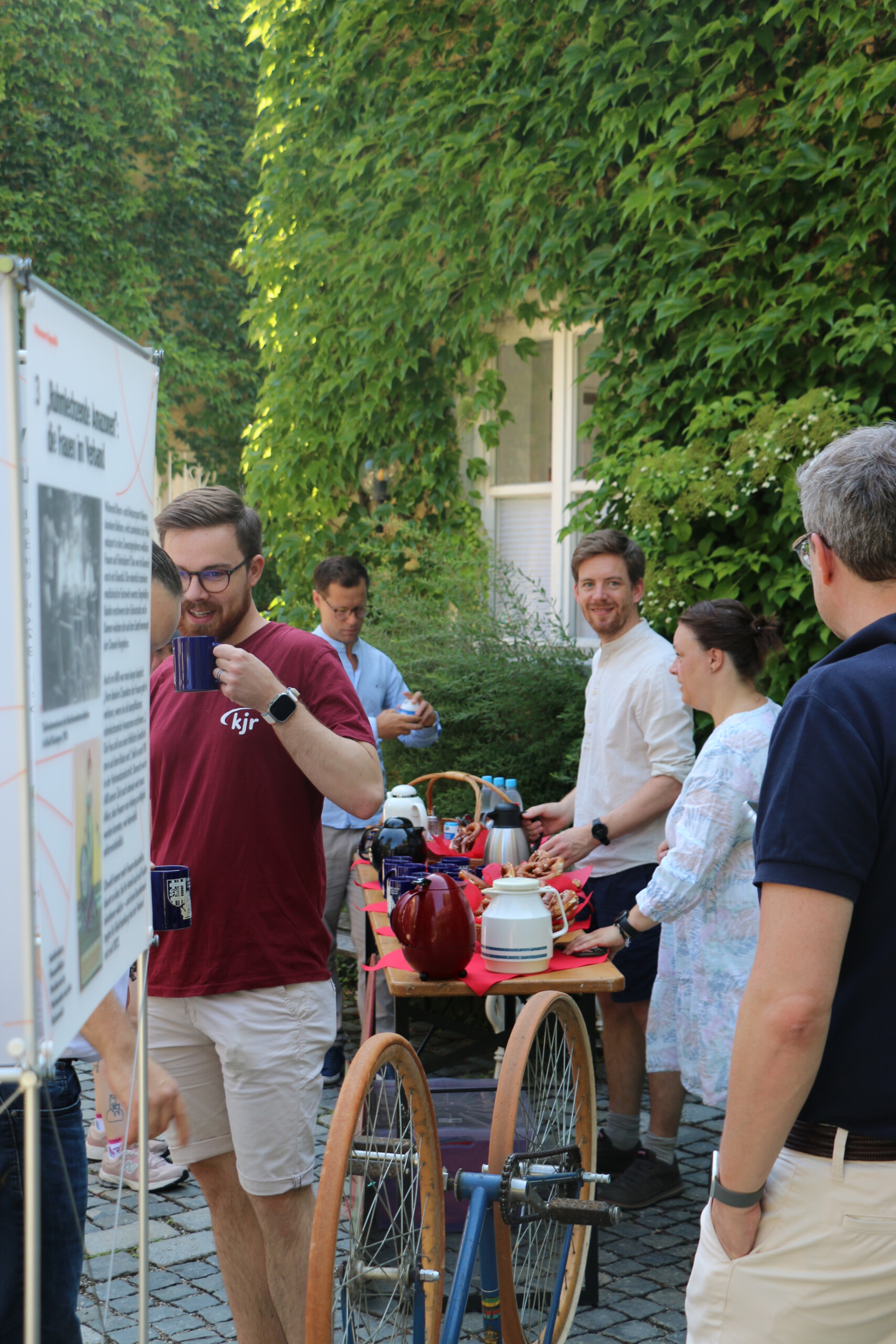 Um einen Biertisch mit Tassen und Kaffeekannen stehen vor einer grün bewachsenen Wand in einem gepflasterten Innenhof mehrere Menschen