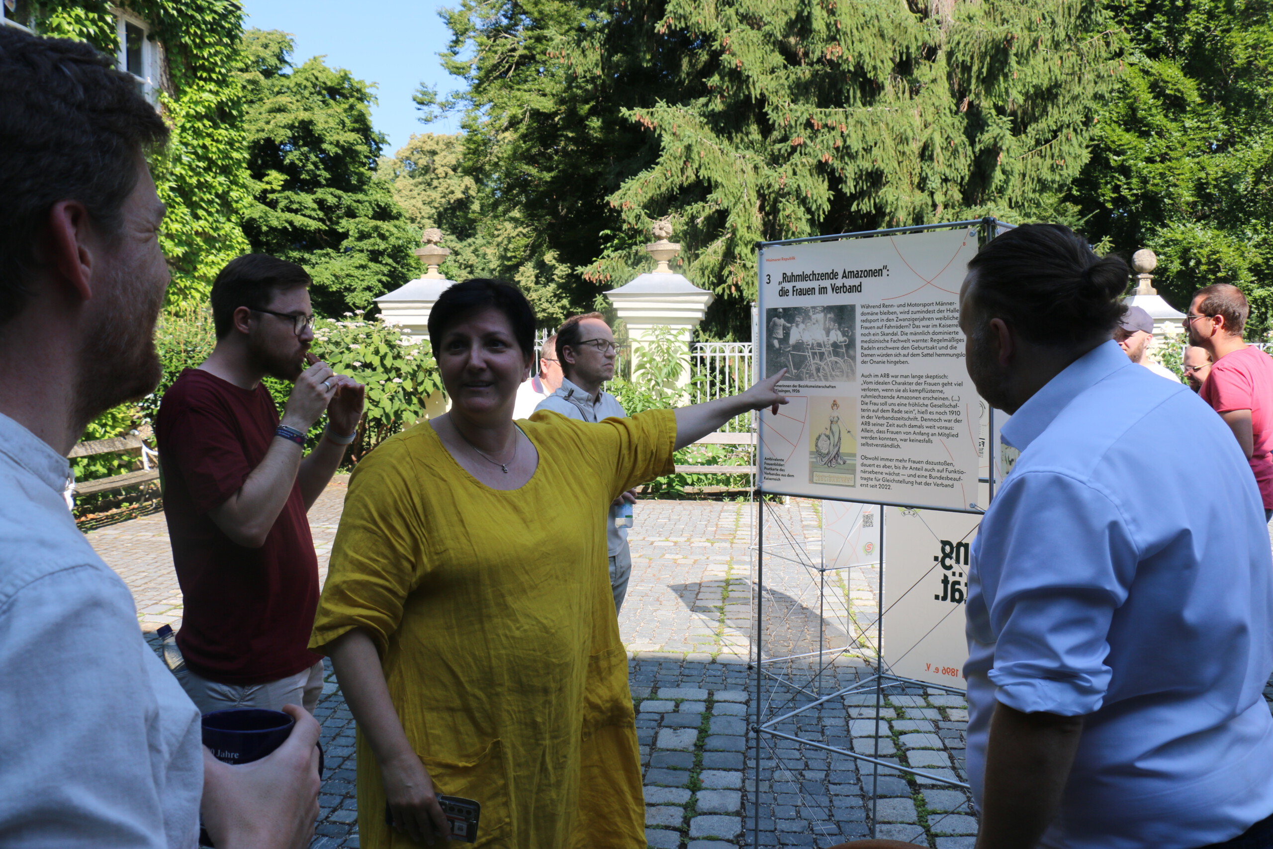 In einem gepflasterten Innenhof stehen mehrere Personen um Ausstellungstafel verteilt, während eine Frau in gelbem Kleid mit ausgestrecktem Arm auf eine Tafel zeigt