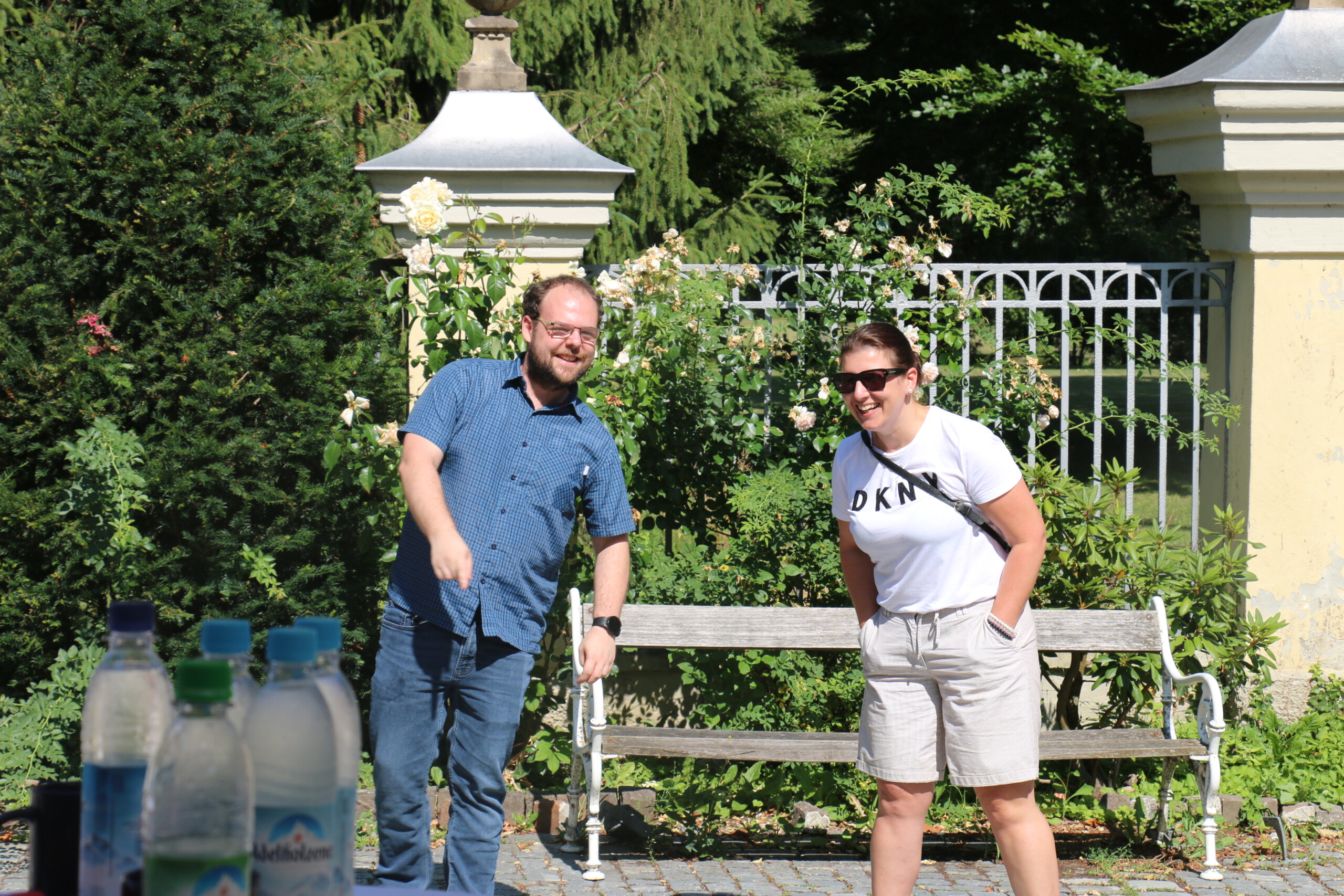 Ein Mann im blaukarierten Kurzarmhemd und eine Frau mit Sonnenbrille im weißen T-Shirt stehen lachend nebeneinander.