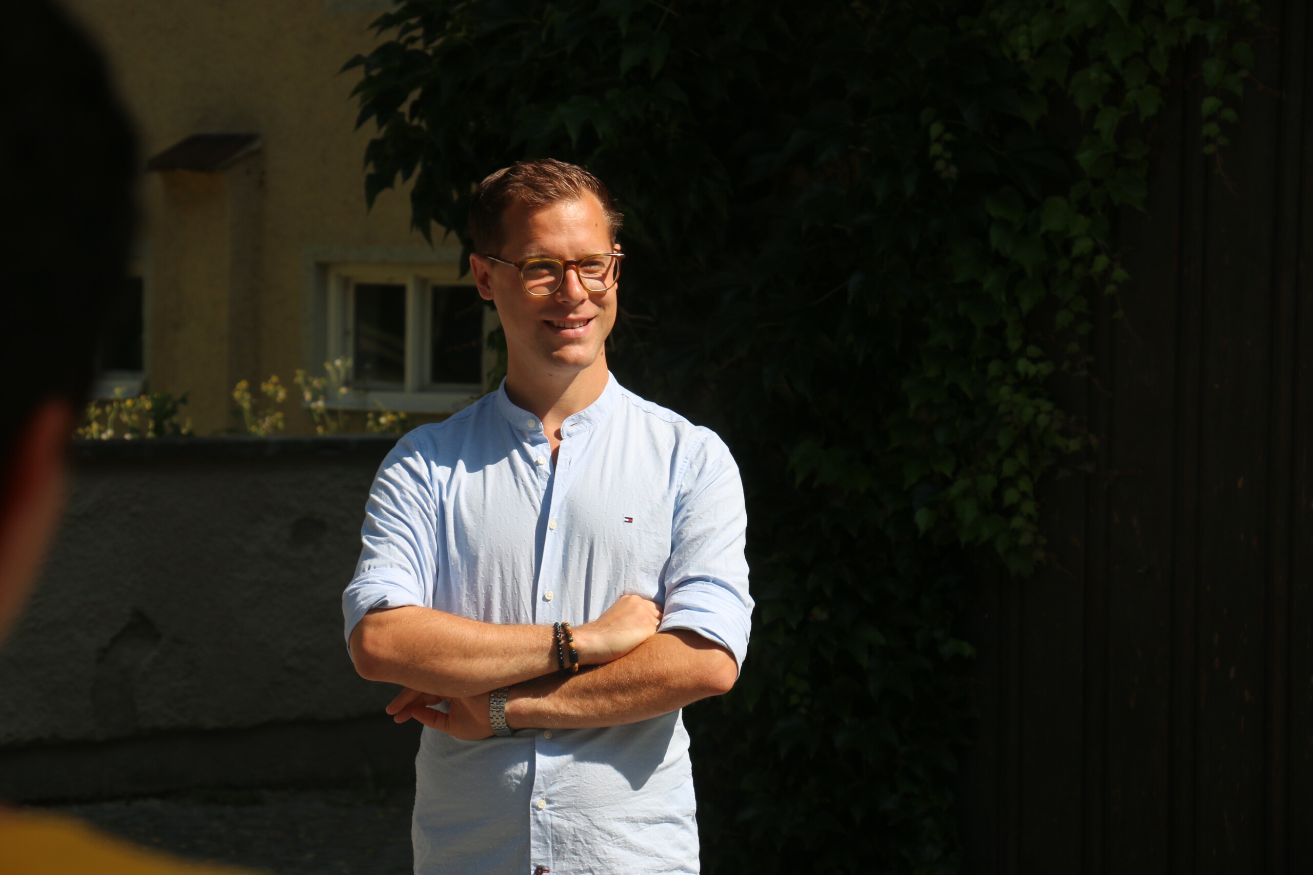 Ein Mann mit Brille und hellblauem Hemd steht mit verschränkten Armen in der Sonne und lächelt