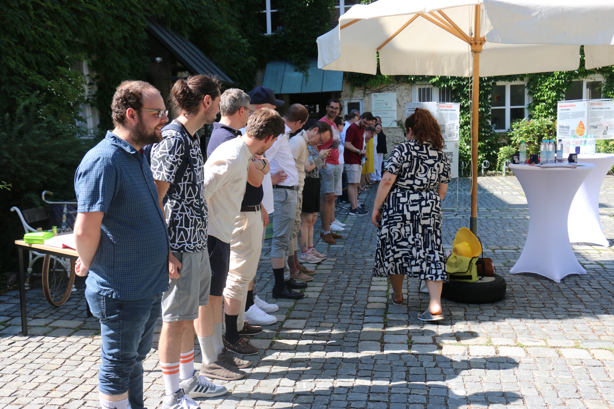 Mehrere Personen stehen aufgereiht in einem gepflasterten Innenhof nebeneinander und haben ihr Hosen hochgekrempelt, damit die Sockenlänge zu erkennen ist. Eine Frau im gemusterten Kleid inspiziert die Socken im vorbeigehen.