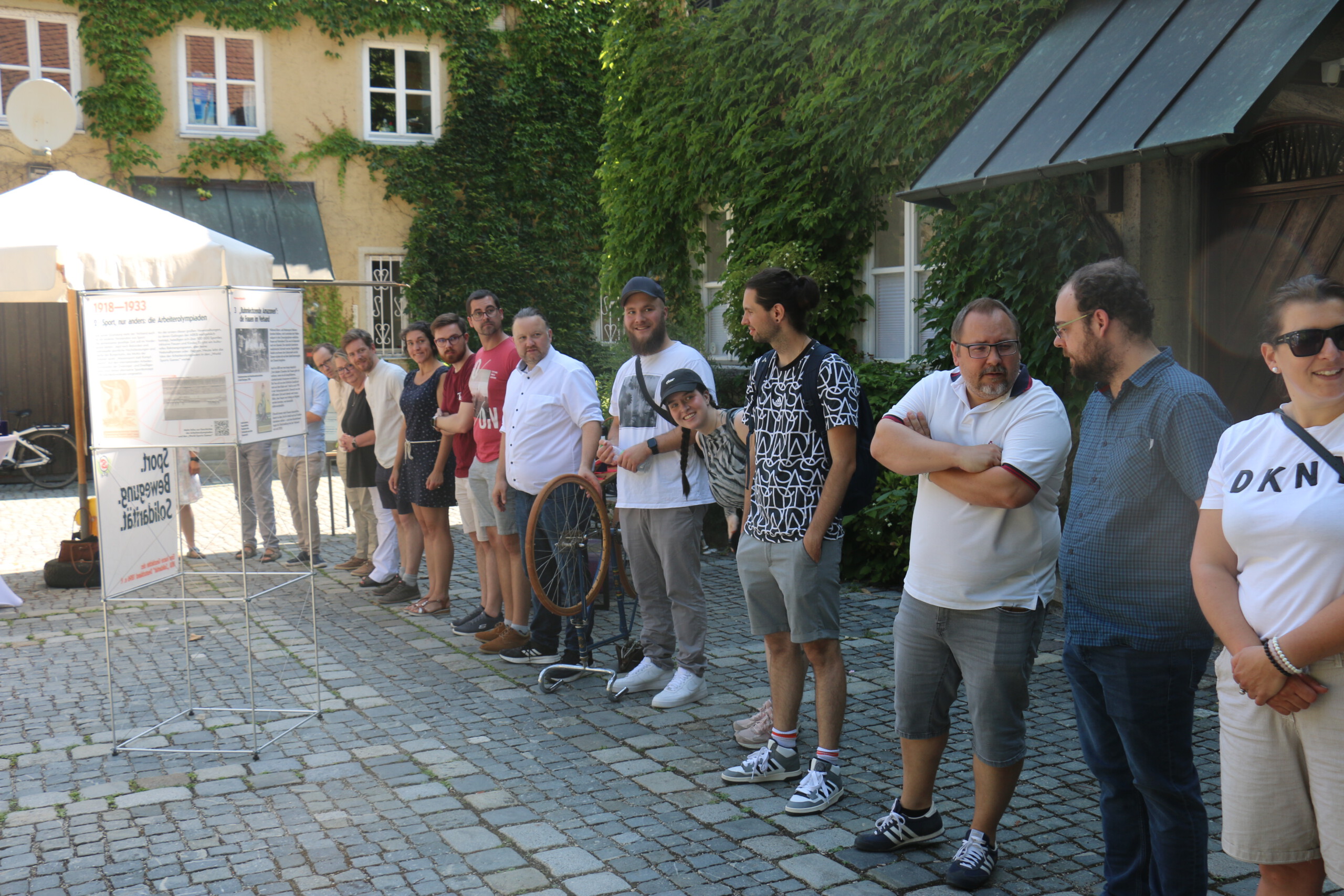 Mehrere Personen stehen in einem gepflasterten Innenhof in einer Reihe nebeneinander und blicken in die Kamerarichtung