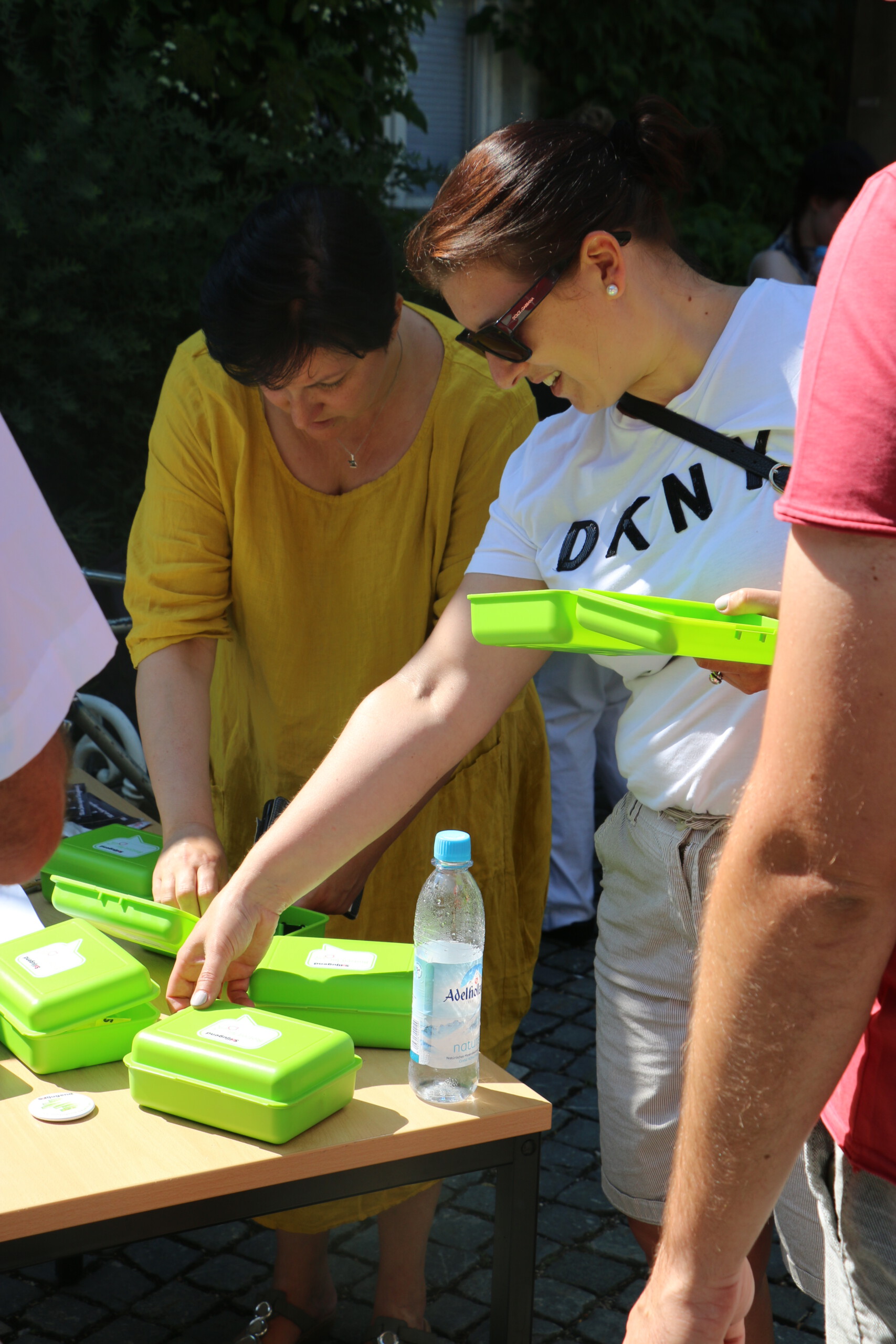 Zwei Frauen stehen an einem Tisch in der Sonne und schauen sich hellgrüne Brotzeitboxen der Solijugend und deren Inhalt an