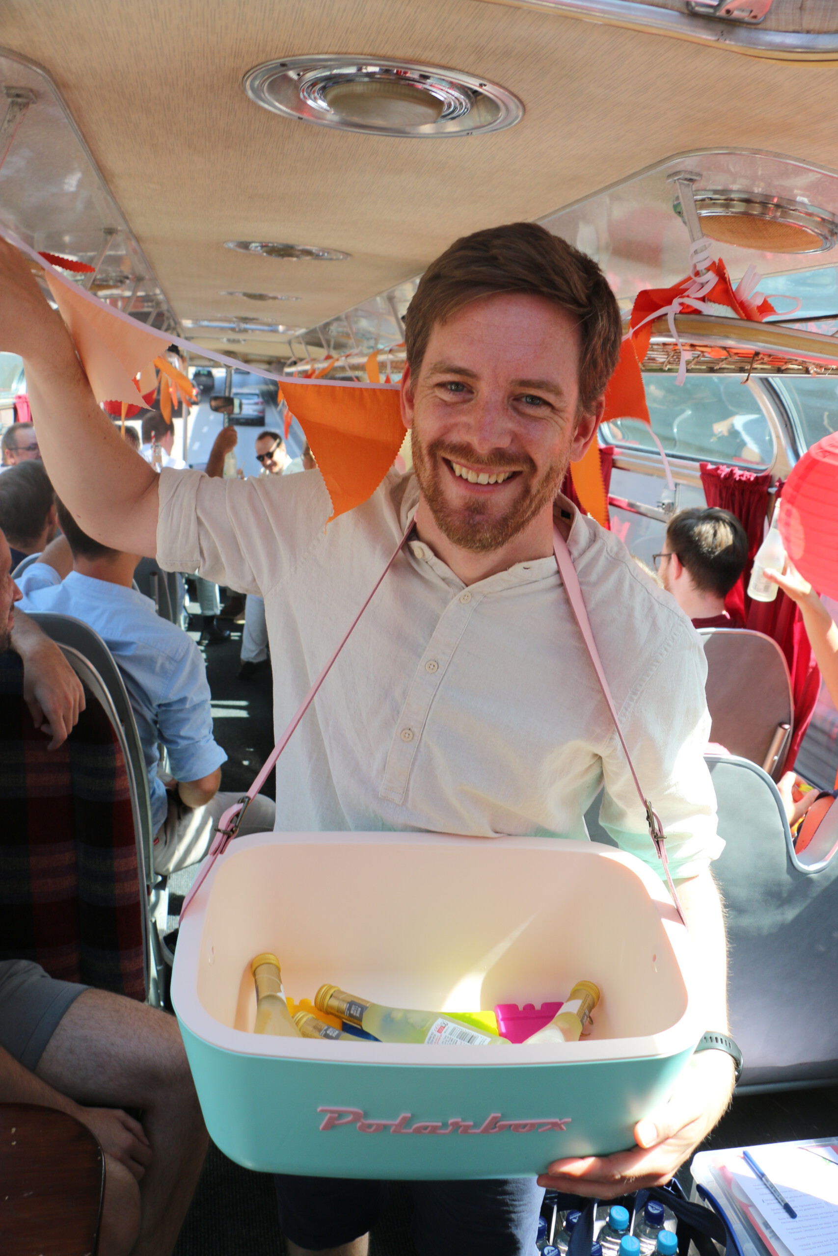 Ein Mann in beigem Leinenhemd posiert lachend mit einer Retro-Kühl-Polarbox gefüllt mit Piccolo-Flaschen im Mittelgang eines Oldtimer-Buses
