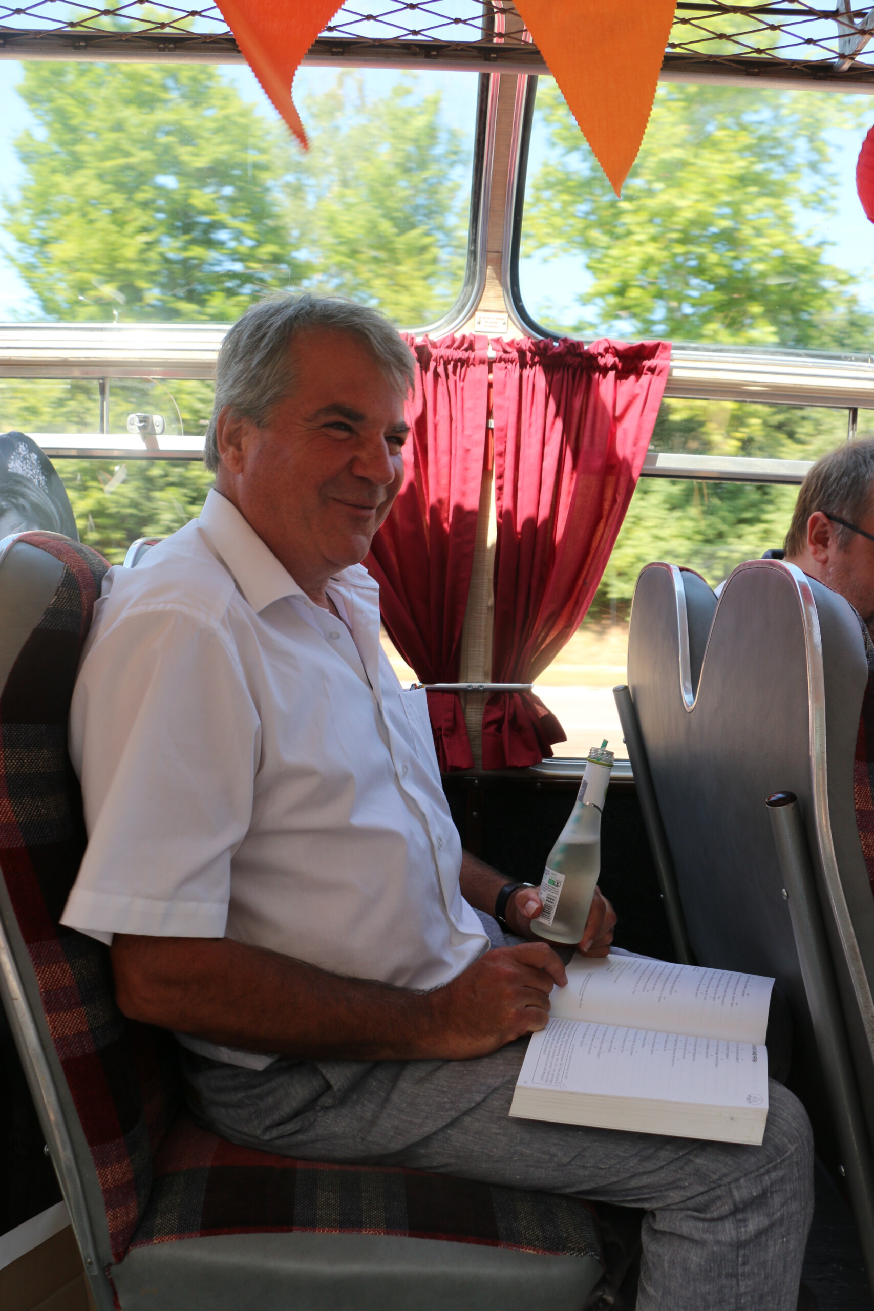 Ein Mann mit weißem Hemd sitzt lachend im Oldtimer-Bus, während er eine Flasche mit Strohhalm und ein Liederbuch in den Händen hält