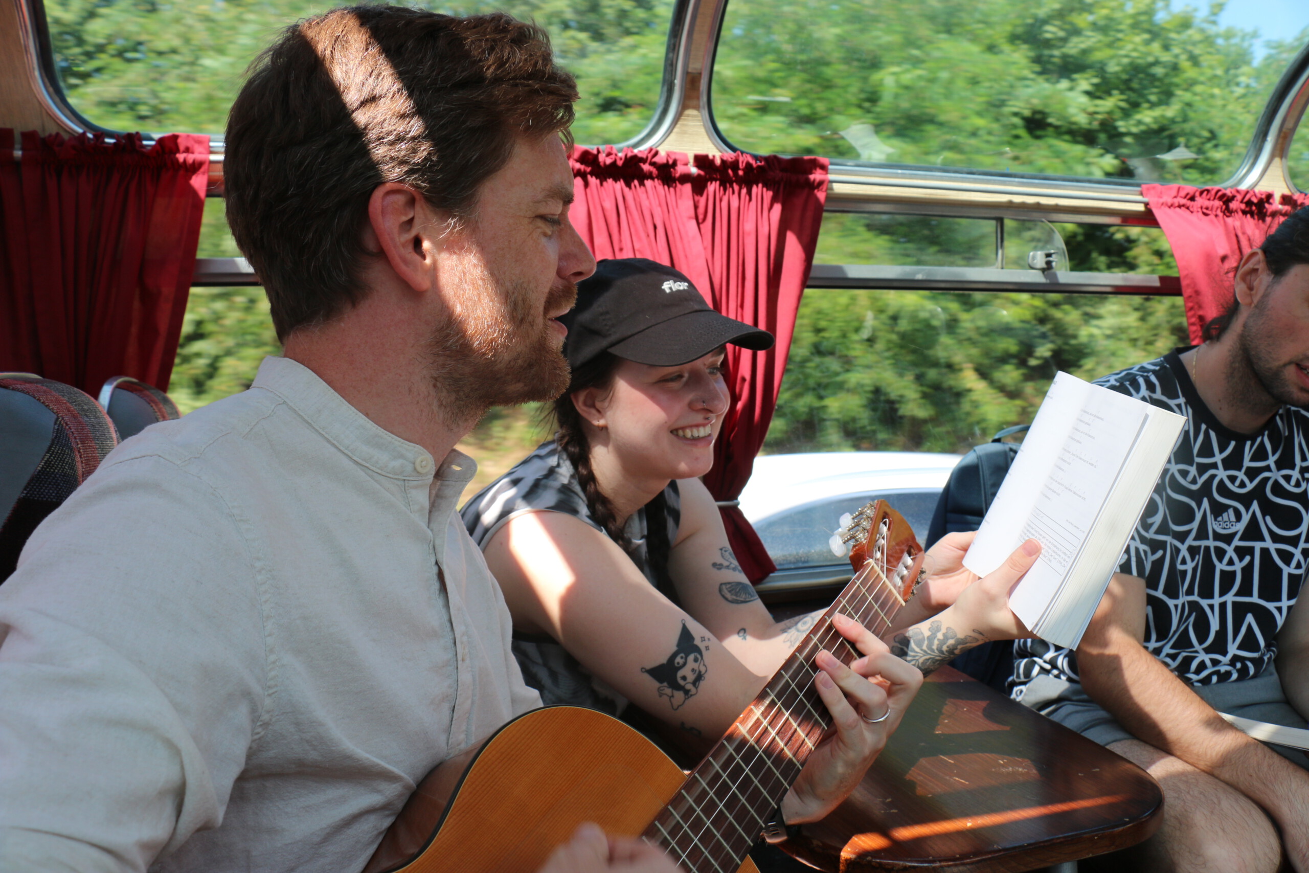 In einem Oldtimer-Bus mit Panoramafenstern spielt ein Mann in beigem Hemd Gitarre, während eine junge Frau mit schwarzen Zöpfen, Cap und Tattoos auf den Armen ihm das Liederbuch hält