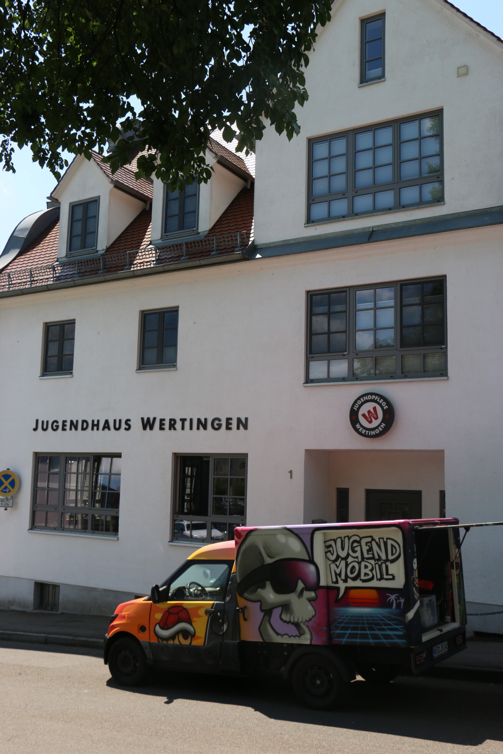 Blick auf das Jugendhaus Wertingen: ein mehrstöckiges weiß gestrichenes Gebäude mit großen Fenstern, davor ein mit Graffiti bemaltes Jugendmobil