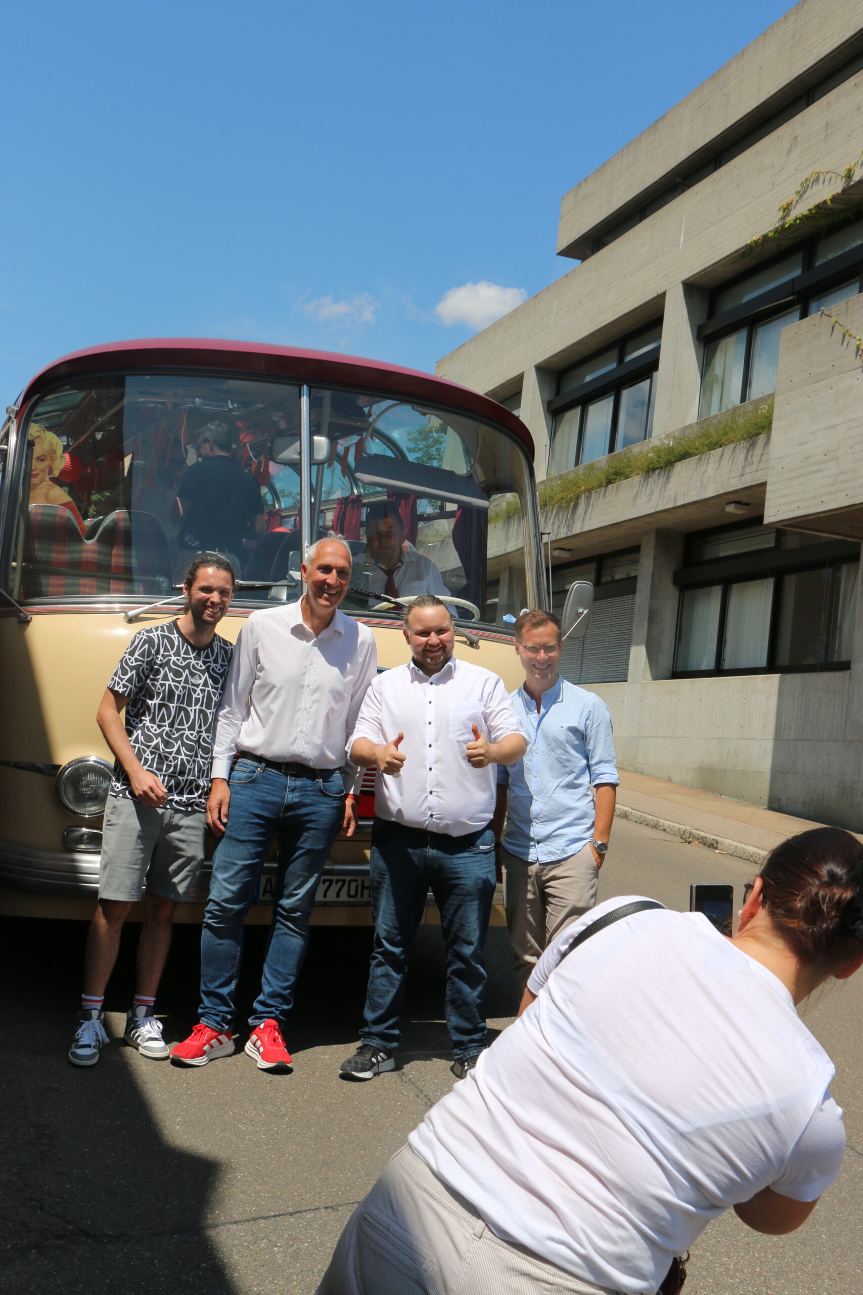 Vor dem rot-beigen Oldtimer-Bus posieren vier Männer, während eine Frau ein Handyfoto von ihnen macht