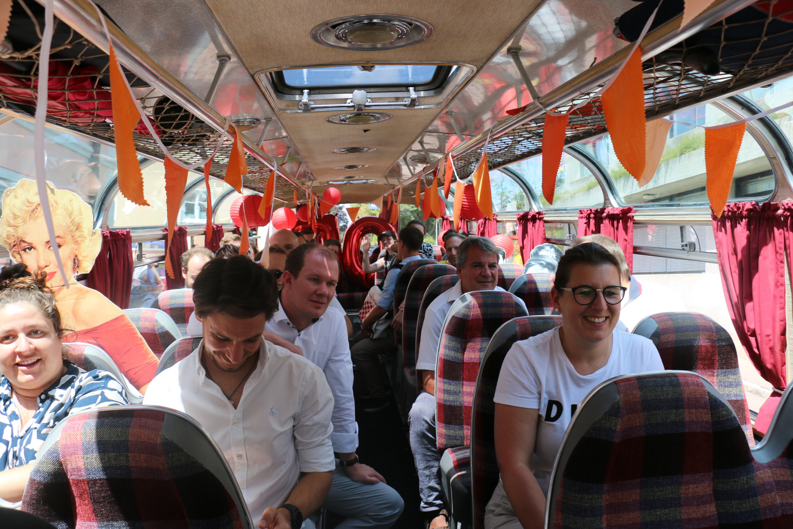 Blick von vorne durch einen Oldtimer-Bus mit rot-grau karierten Sitzpolstern und orange-roter Dekoration: Personen auf den Sitzen unterhalten sich und lachen in die Kamera