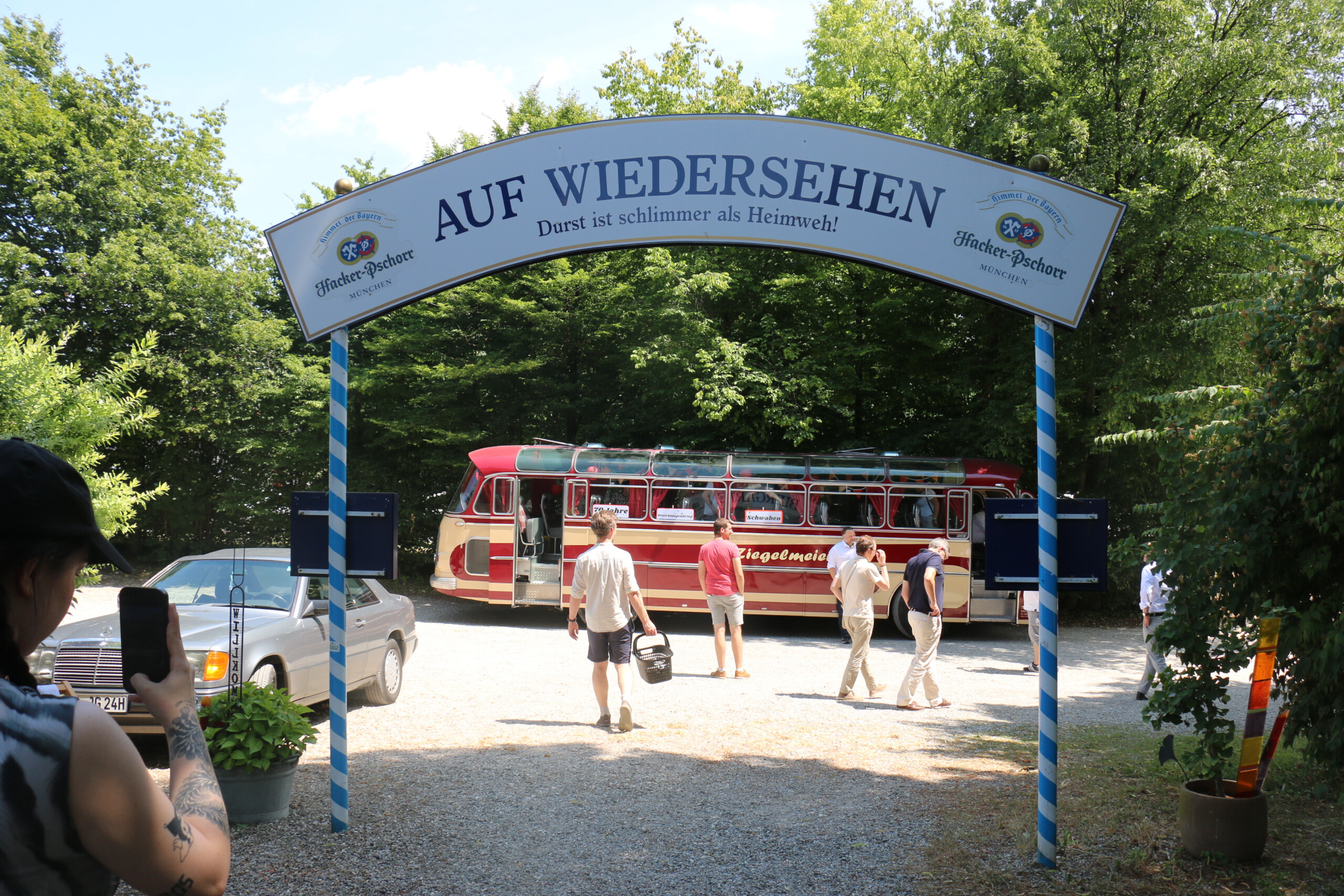 Im Vordergrund der Ausgang eines Biergartens mit einem Bogen, auf dem steht: Auf Wiedersehen. Durst ist schlimmer als Heimweh! Im Hintergrund vereinzelt Personen, die auf einen rot-beigen Oldtimer-Bus zugehen