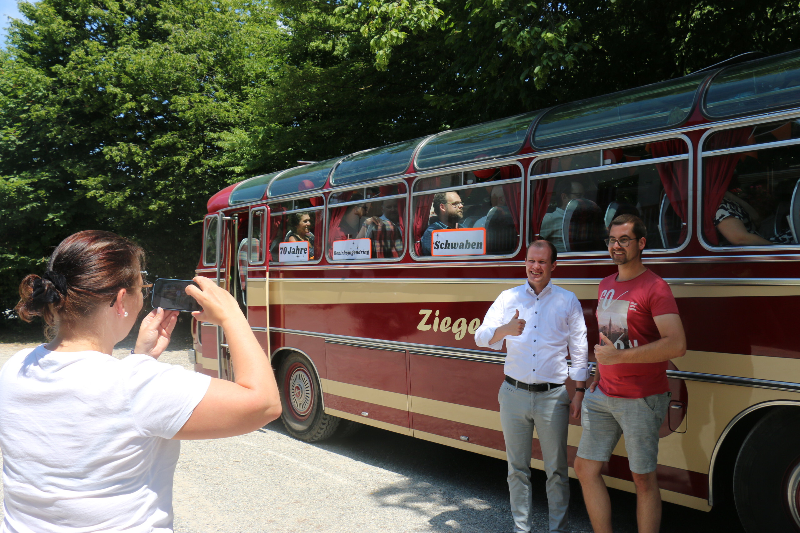 Zwei Männer posieren mit Daumen hoch vor einem rot-beigem Oldtimer-Bus, während eine Frau in weißem T-Shirt ein Handyfoto von ihnen macht