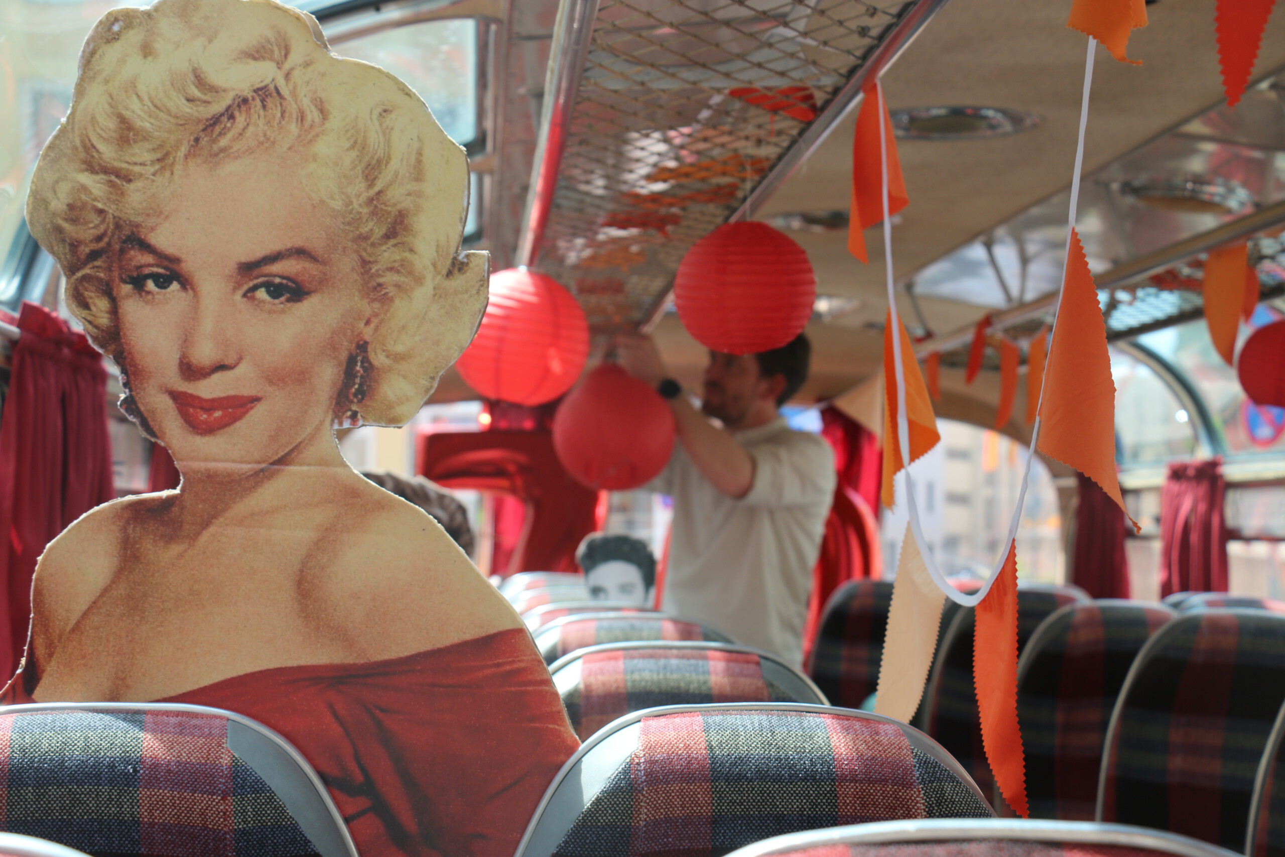 Blick im Oldtimer-Bus, im Vordergrund eine Pappfigur von Marilyn Monroe, im Hintergrund ein Mann in beigem Hemd, der die Dekoration abhängt