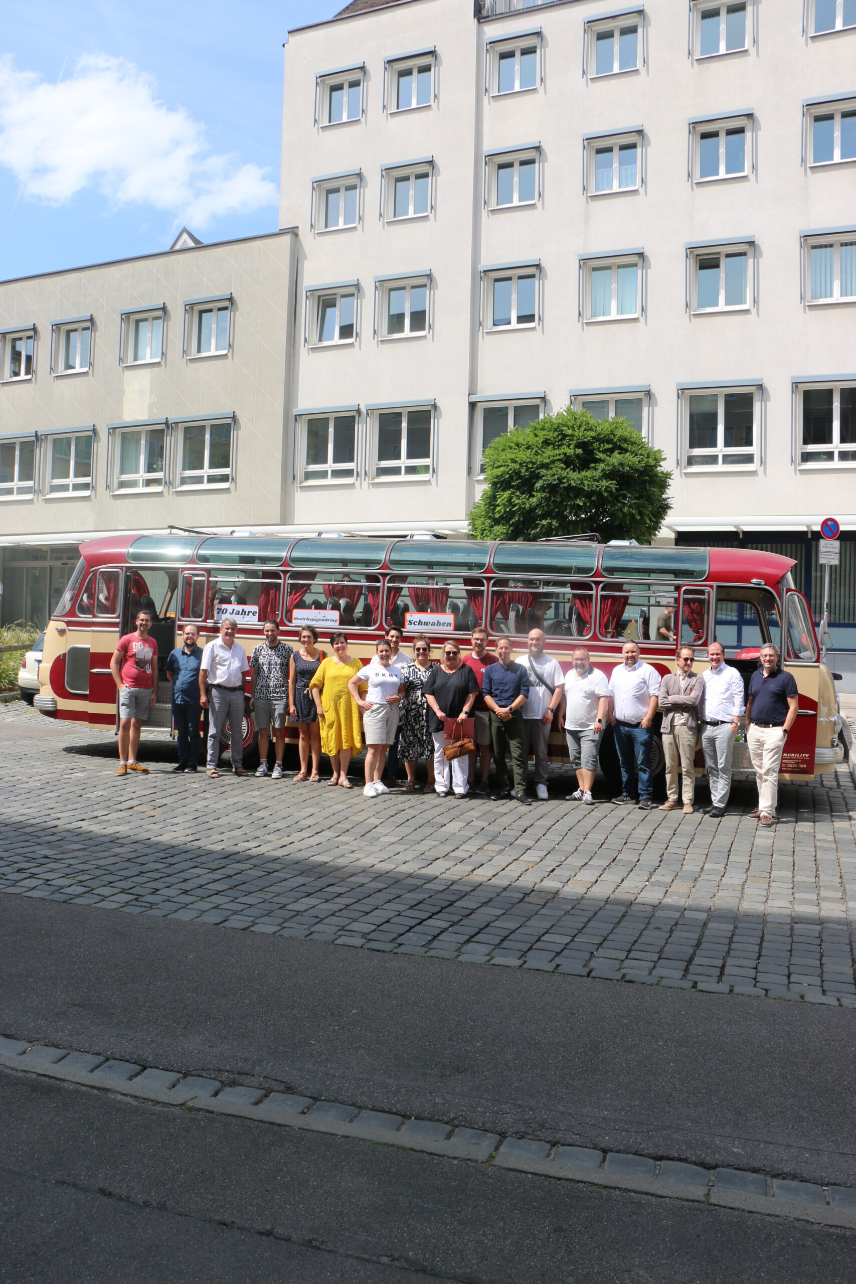 Vor einem rot-beigem Oldtimer-Bus posieren 19 Personen aufgereiht bei Sonnenschein