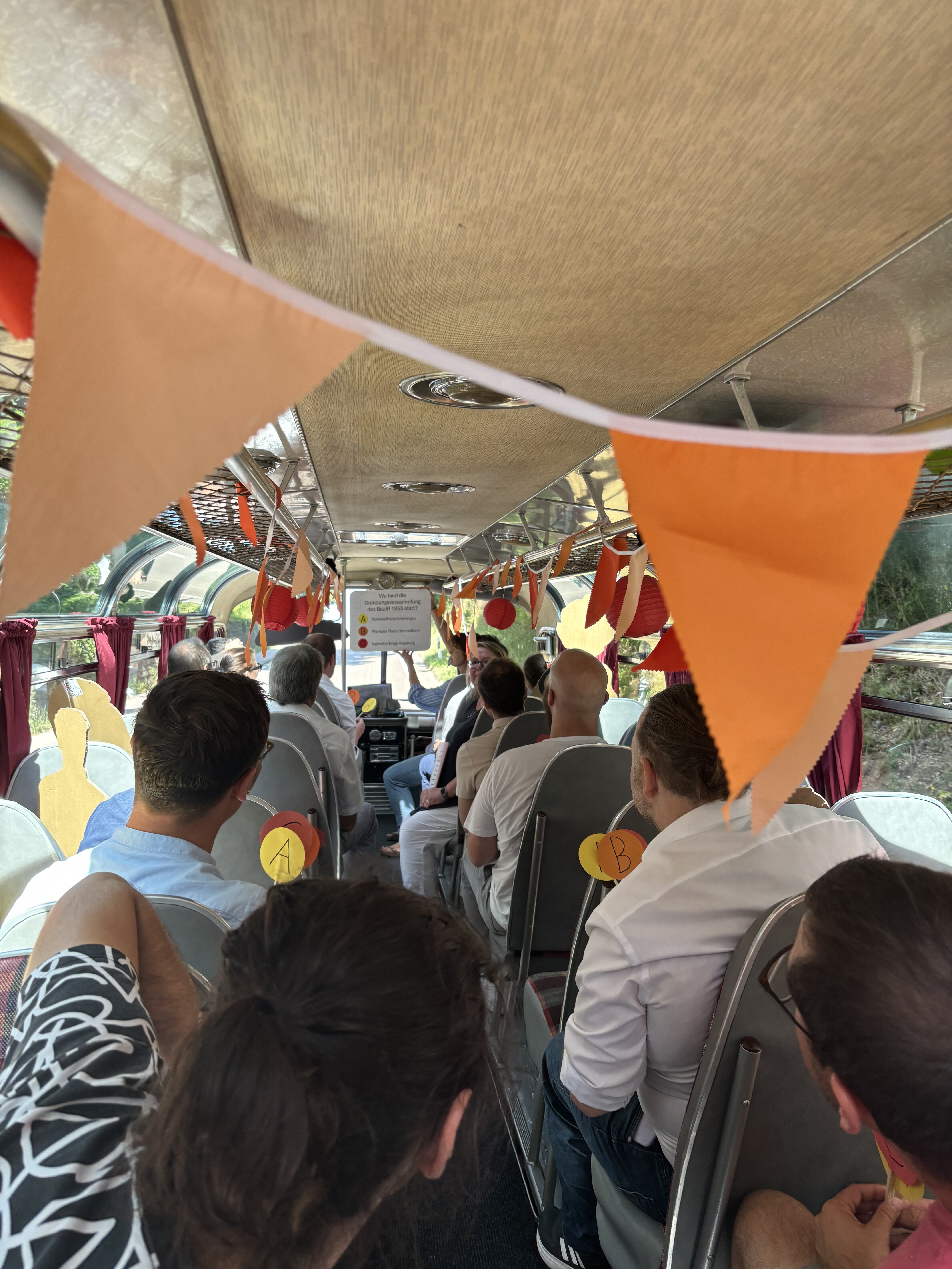 Blick von den hinteren Sitzreihen eines Oldtimer-Buses nach vorne. Vorne wird ein Schild hochgehoben, die sitzenden Menschen haben drei Schilder mit A,B und C in der Hand als Antwortmöglichkeiten